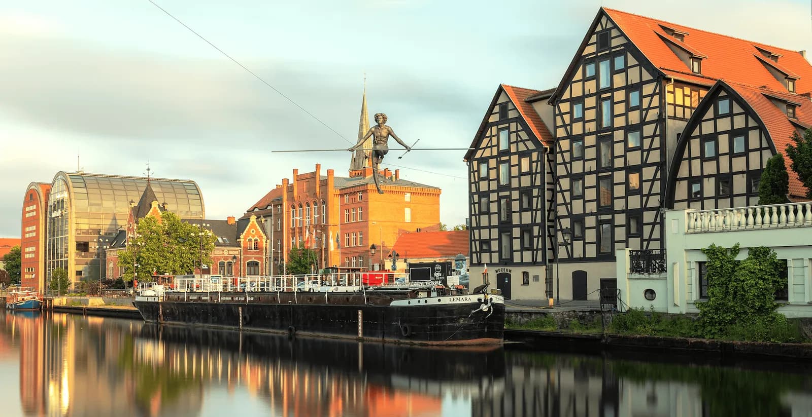 A bronze statue of a tightrope walker, suspended high above a canal, is the focal point of a cityscape featuring historic buildings with timber framing and red roofs, as well as a large barge floating on the water.